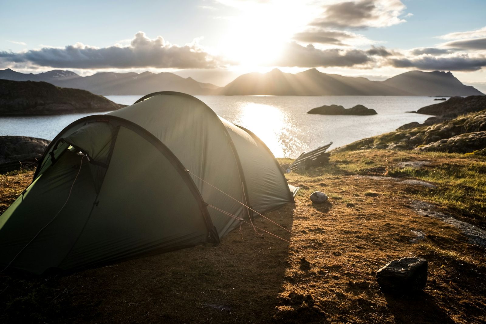 A low tent pitched on a rocky shoreline at sunrise, with mist over a lake and mountains behind.